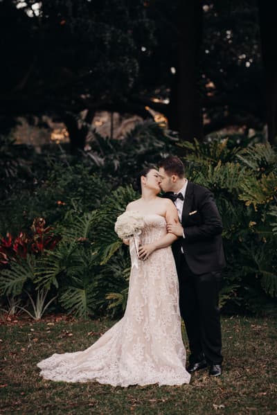 The bride Aria and groom Antony share a kiss during their couple portraits session at City Botanic Gardens. Aria holds a bouquet of white flowers and wears a strapless lace wedding gown, while Antony is dressed in a black tuxedo with a bow tie. Brisbane wedding photographers by She Said Yes.