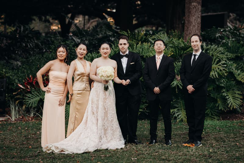Aria the bride in a white lace gown holding a bouquet of white roses stands next to Antony the groom in a black tuxedo with a bow tie, flanked by two bridesmaids in gold dresses on the left and two groomsmen in black suits on the right, posing for a group portrait at City Botanic Gardens.
