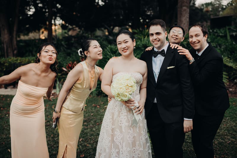 The bride Aria in a lace wedding gown holding a bouquet of white roses stands next to the groom Antony in a black tuxedo with a bow tie, accompanied by two women in formal dresses and one man in a black suit, posing outdoors at City Botanic Gardens. Best Brisbane wedding photographer by She Said Yes.