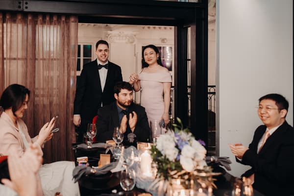 Aria and Antony stand holding hands behind a seated guest who is clapping at a reception table in Donna Chang, while other guests sit around the table, one using a phone and another clapping.