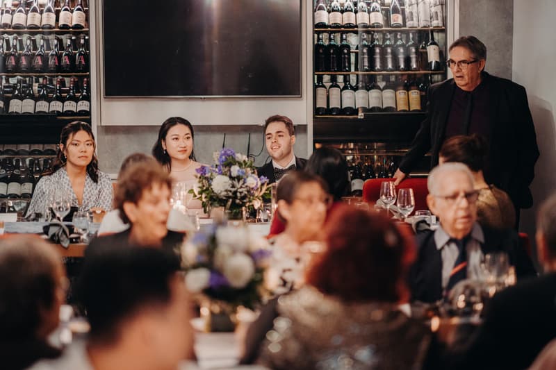 Aria and Antony sit at a head table with two other guests at Donna Chang during the wedding reception, with wine bottles displayed behind them and guests seated at tables in the foreground.