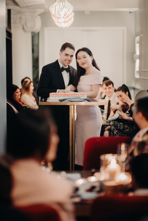 Aria and Antony stand together behind a table with a cake at the reception stage in Donna Chang, surrounded by seated guests.