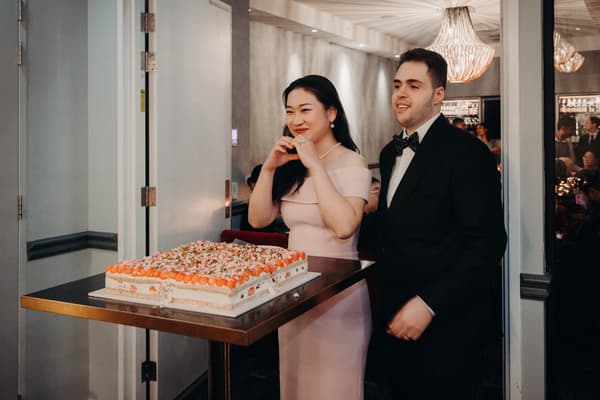 The bride Aria and groom Antony stand together behind a table with a large rectangular cake at the reception stage in Donna Chang. Aria forms a heart shape with her hands while Antony stands beside her in a black suit and bow tie.