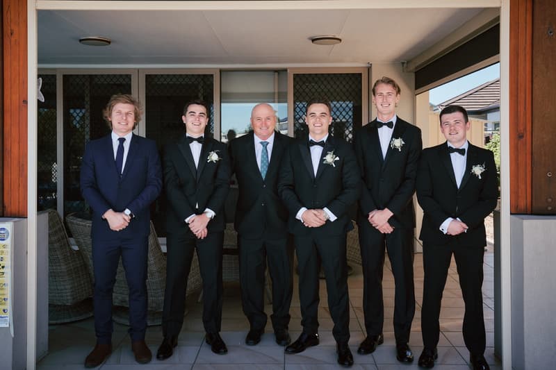 James the groom stands with five men, including groomsmen in tuxedos with boutonnieres and an older man in a suit, posing outside a building at Sandstone Point Hotel.