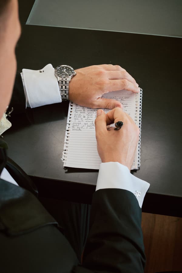 The groom at Sandstone Point Hotel writes notes on a spiral notebook while seated at a dark table, wearing a black suit with white shirt cuffs and a silver wristwatch.