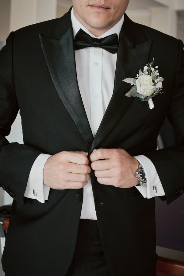The groom adjusts his black tuxedo jacket, wearing a white shirt with cufflinks and a black bow tie, with a white rose boutonniere pinned to his lapel.