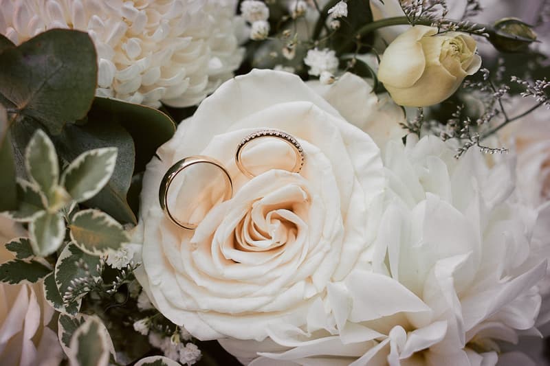 Two wedding rings placed on a white rose surrounded by other white flowers and greenery.