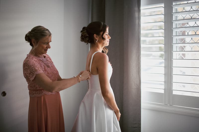 The mother of the bride helps Ashleigh, the bride, by fastening the back of her wedding dress near a window with white shutters.