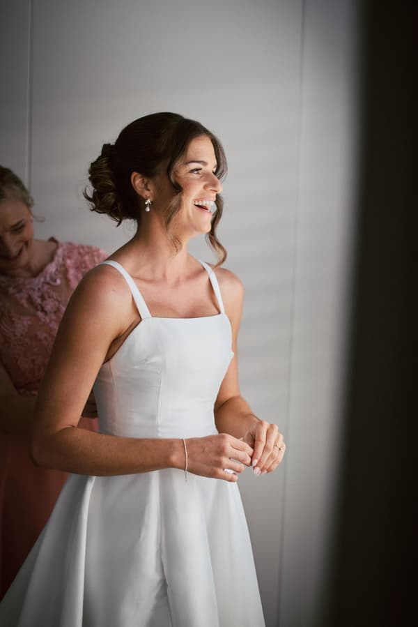 The bride Ashleigh stands smiling in a white wedding dress while an older woman, likely the mother of the bride, adjusts the back of her dress at Sandstone Point Hotel.