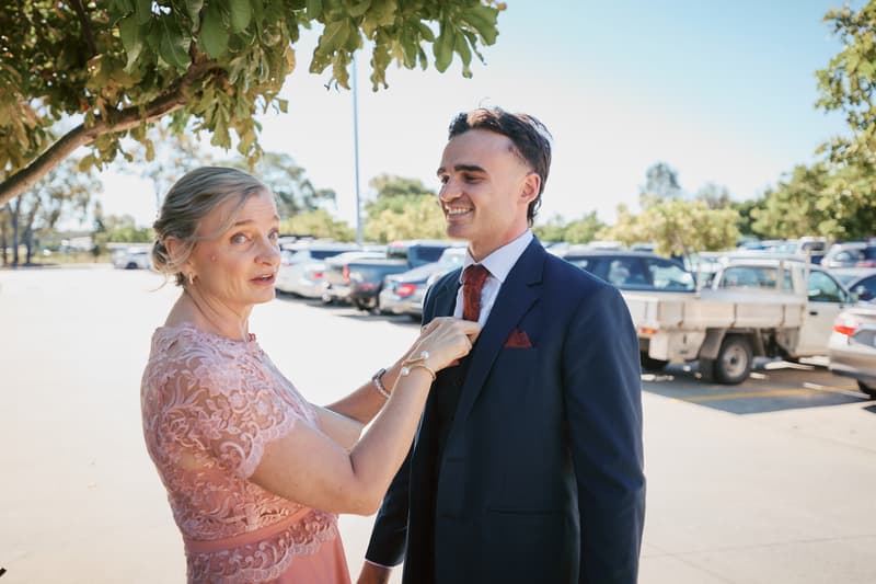 The groom James stands outdoors in a parking area at Sandstone Point Hotel — The Pavilion while an older woman, likely the groom's mother, adjusts his tie.