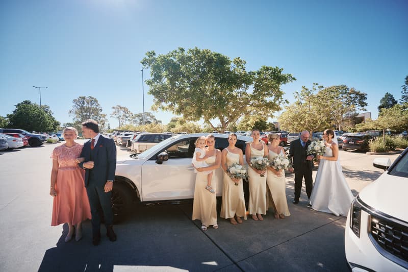 At Sandstone Point Hotel — The Pavilion parking area, the bride Ashleigh in a white gown stands with an older man, while the groom James in a dark suit stands arm-in-arm with an older woman in a pink dress. Four bridesmaids in matching cream dresses hold bouquets, one holding a young child.