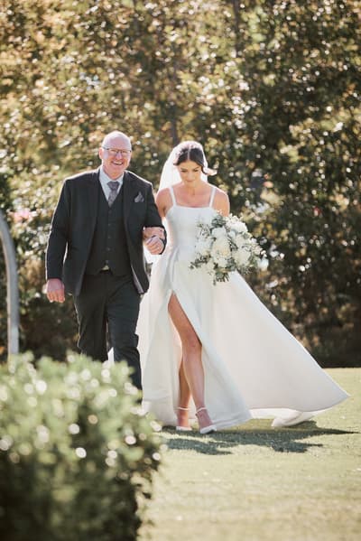 The bride Ashleigh walks arm-in-arm with an older man, likely her father, outdoors, holding a bouquet of white flowers. Best time of year to get married by She Said Yes.