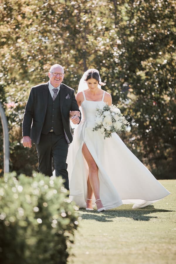 The bride Ashleigh walks arm-in-arm with an older man, likely her father, outdoors at Sandstone Point Hotel — The Pavilion, holding a bouquet of white flowers.