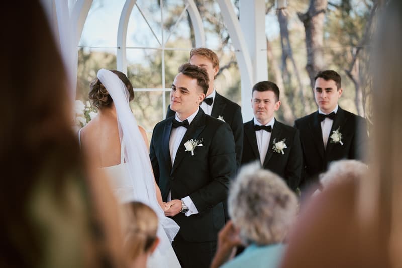 James and Ashleigh stand facing each other at the altar during their wedding ceremony at Sandstone Point Hotel — The Pavilion, with groomsmen standing behind James and guests partially visible in the foreground.