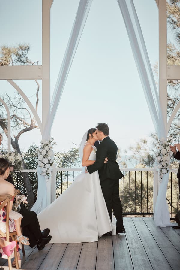 Bride Ashleigh and groom James share a kiss at the altar during their wedding ceremony at Sandstone Point Hotel — The Pavilion, surrounded by floral decorations and seated guests.