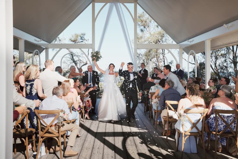 Bride Ashleigh and groom James walk down the aisle holding hands at Sandstone Point Hotel — The Pavilion, as guests seated on both sides throw confetti.