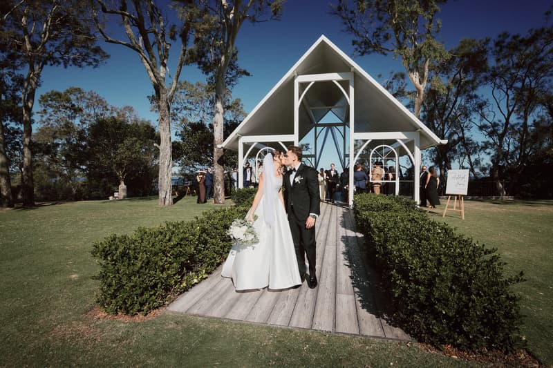 Bride Ashleigh and groom James kiss at the Sandstone Point Hotel — The Pavilion after their wedding ceremony, with guests visible in the background near the altar structure.