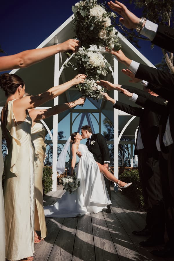 Bride Ashleigh and groom James share a kiss under a white wooden arch at Sandstone Point Hotel, surrounded by bridesmaids in cream dresses and groomsmen in black suits holding bouquets overhead.