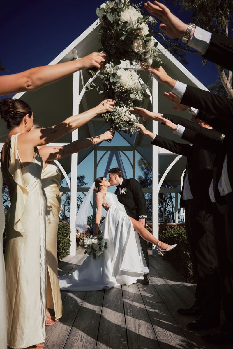 Bride Ashleigh and groom James share a kiss under a white wooden arch, surrounded by bridesmaids in cream dresses and groomsmen in black suits holding bouquets overhead. Brisbane wedding photography by She Said Yes.