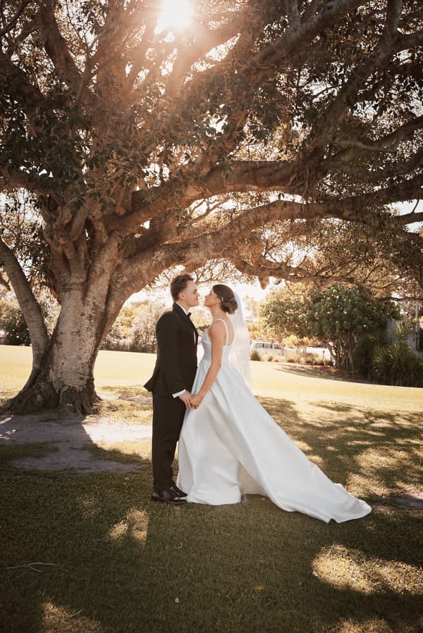 The bride Ashleigh and groom James stand holding hands and facing each other under a large tree at Sandstone Point Hotel during their couple portraits session.