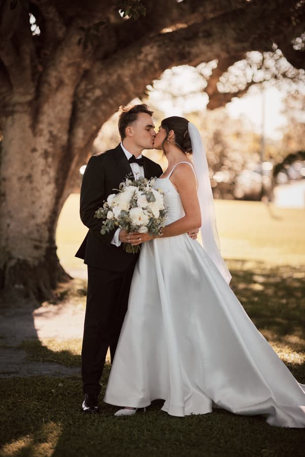 Bride Ashleigh and groom James share a kiss while standing under a large tree at Sandstone Point Hotel. Ashleigh wears a white wedding gown and veil, holding a bouquet of white flowers, while James wears a black tuxedo with a bow tie.