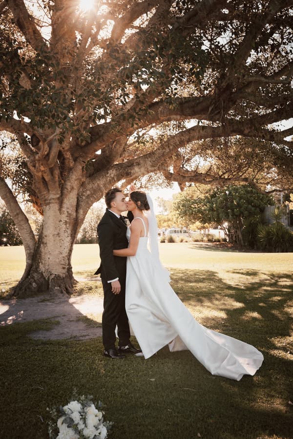 The bride Ashleigh and groom James share a kiss under a large tree at Sandstone Point Hotel during their couple portraits session. Ashleigh wears a white wedding gown with a long train and veil, while James is dressed in a black suit. A bridal bouquet lies on the grass nearby.