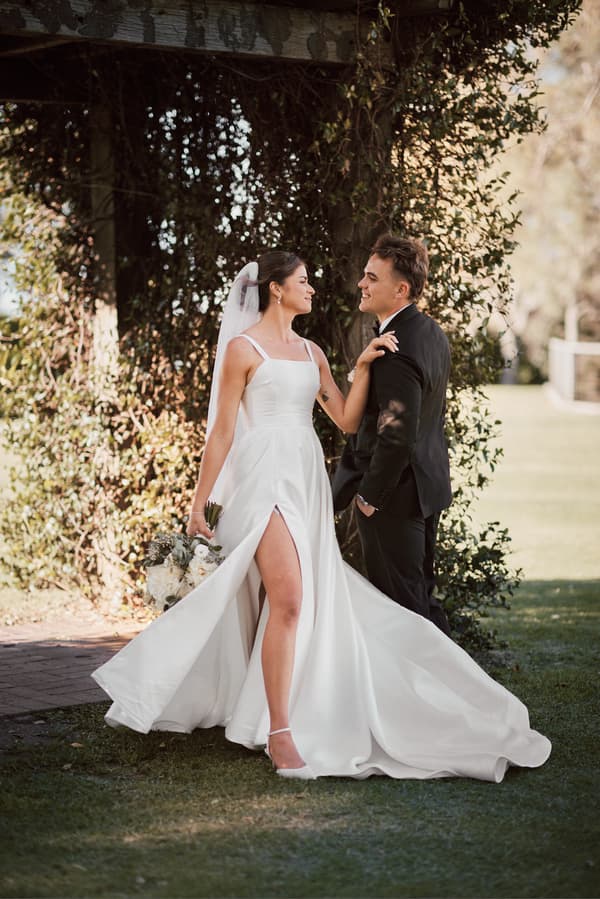 The bride Ashleigh in a white wedding gown with a high slit and veil holds a bouquet and faces the groom James, who is dressed in a black suit, standing under a leafy arch at Sandstone Point Hotel.