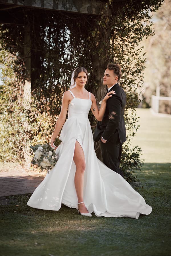 The bride Ashleigh in a white wedding gown holding a bouquet stands next to the groom James in a black suit under a leafy arch at Sandstone Point Hotel.