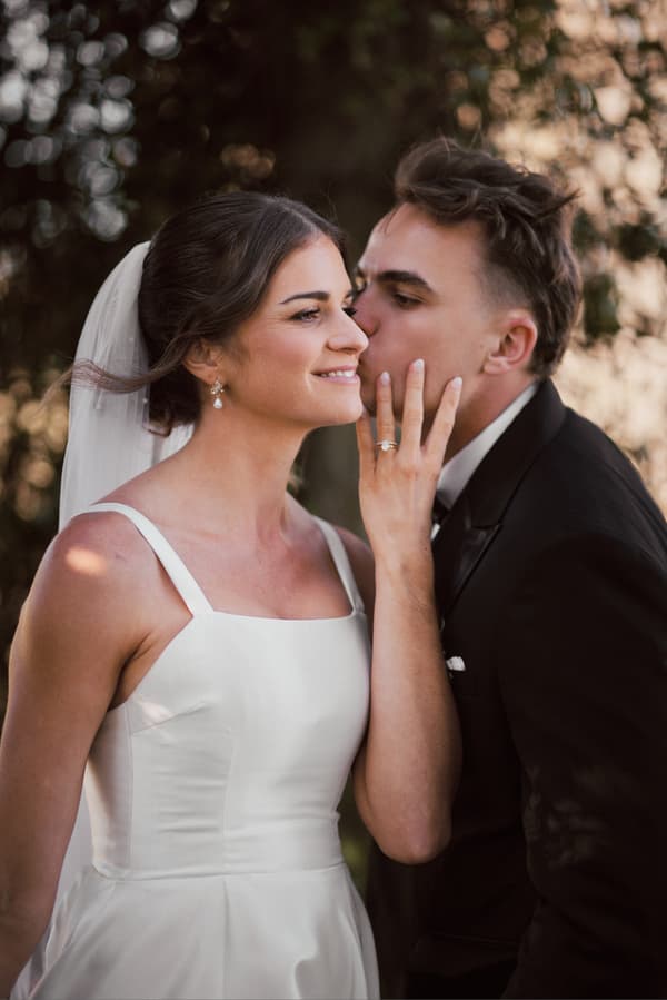 The bride Ashleigh in a white wedding dress and veil smiles as the groom James in a black suit kisses her cheek at Sandstone Point Hotel.