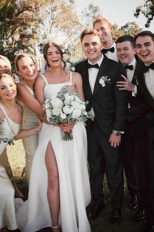 Bride Ashleigh in a white wedding dress holding a bouquet of white flowers stands next to groom James in a black tuxedo with a bow tie, surrounded by bridesmaids in light-colored dresses and groomsmen in black tuxedos at Sandstone Point Hotel outdoors.