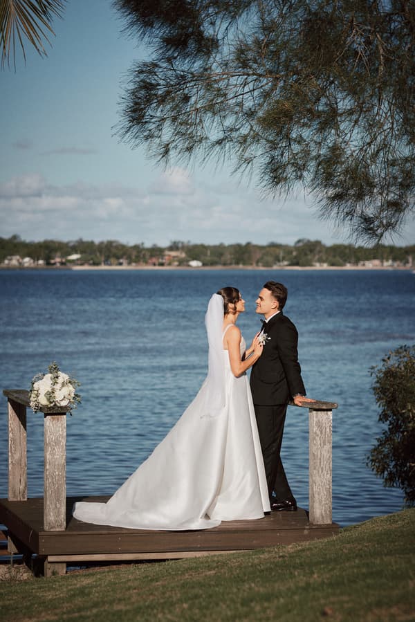 The bride Ashleigh in a white wedding gown and veil stands facing the groom James in a black suit on a wooden platform by the water at Sandstone Point Hotel. A bouquet of white flowers is placed on a post to the left, and trees frame the scene.