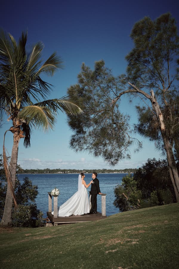 The bride Ashleigh and groom James stand on a small wooden platform by the water at Sandstone Point Hotel, holding hands and facing each other with trees and a clear blue sky in the background.