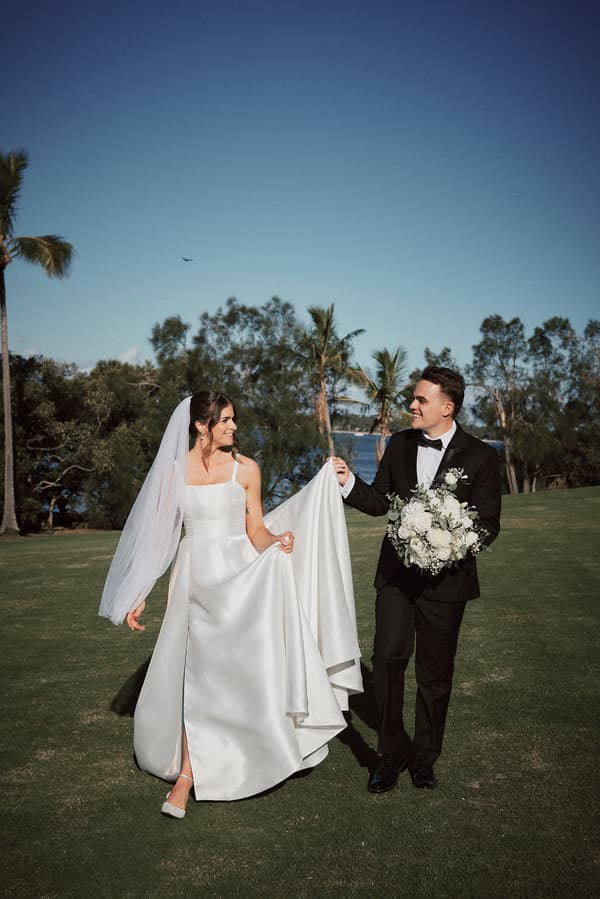 The bride Ashleigh in a white wedding gown and veil walks on grass holding the groom James's hand, who is dressed in a black tuxedo and holding a bouquet of white flowers, at Sandstone Point Hotel outdoors with trees and blue sky in the background.