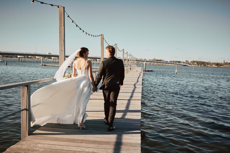 The bride Ashleigh and groom James walk hand-in-hand along a wooden pier at Sandstone Point Hotel, with the bride's veil and dress flowing behind her and a body of water surrounding the pier.