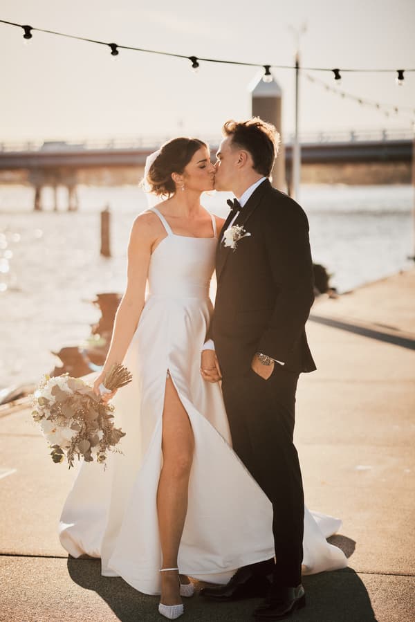 The bride Ashleigh and groom James share a kiss while holding hands at Sandstone Point Hotel, with the bride holding a bouquet and a waterfront background.