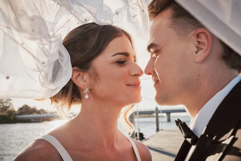 Bride Ashleigh and groom James face each other closely with Ashleigh's veil flowing above them at Sandstone Point Hotel near a waterfront pier.