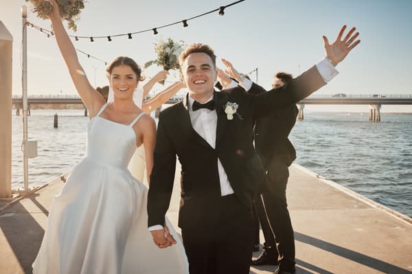 The bride Ashleigh and groom James hold hands and smile at the camera on a pier, with two groomsmen behind them holding bouquets and raising their arms. Destination wedding photographer by She Said Yes.
