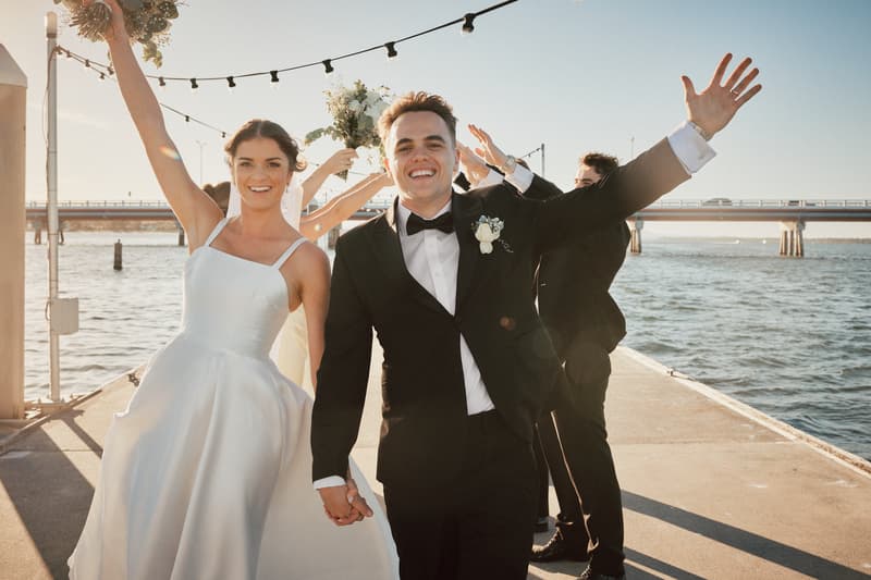 The bride Ashleigh and groom James hold hands and smile at the camera on a pier at Sandstone Point Hotel, with two groomsmen behind them holding bouquets and raising their arms.