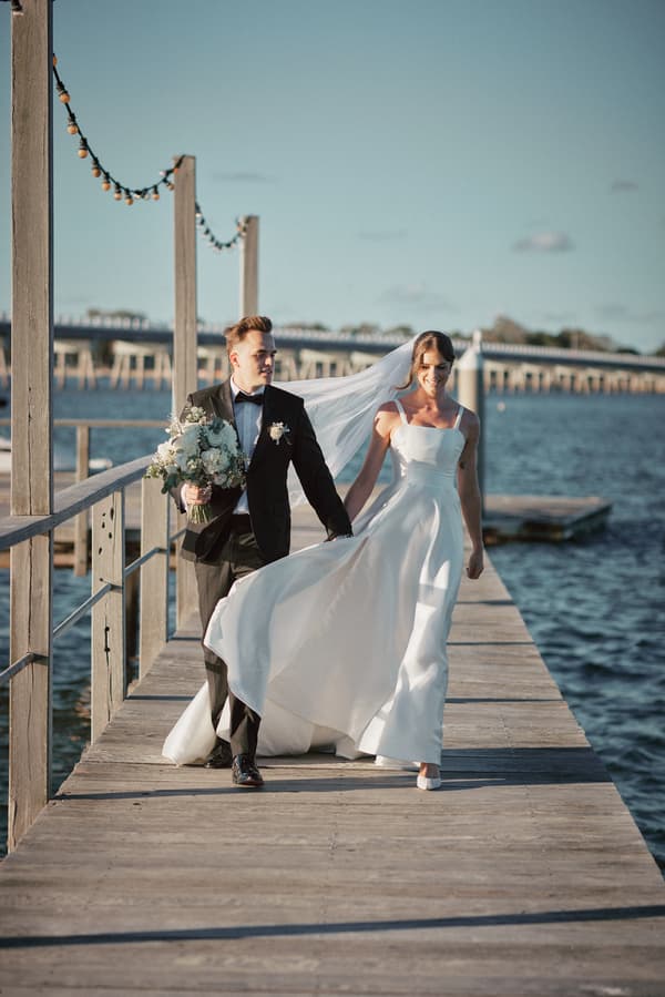 The bride Ashleigh in a white wedding gown and veil walks hand-in-hand with the groom James in a black tuxedo holding a bouquet on a wooden pier at Sandstone Point Hotel with water and a bridge in the background.