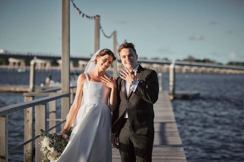 The bride Ashleigh in a white wedding gown holding a bouquet and the groom James in a black suit stand together on a wooden pier at Sandstone Point Hotel, both showing their wedding rings to the camera.