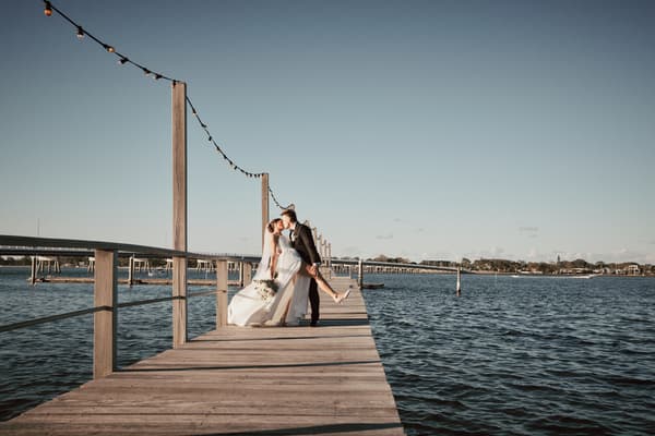 The bride Ashleigh and groom James share a kiss on a wooden pier, with the bride holding a bouquet and the water surrounding them under a clear sky. Gold Coast wedding videographer by She Said Yes.