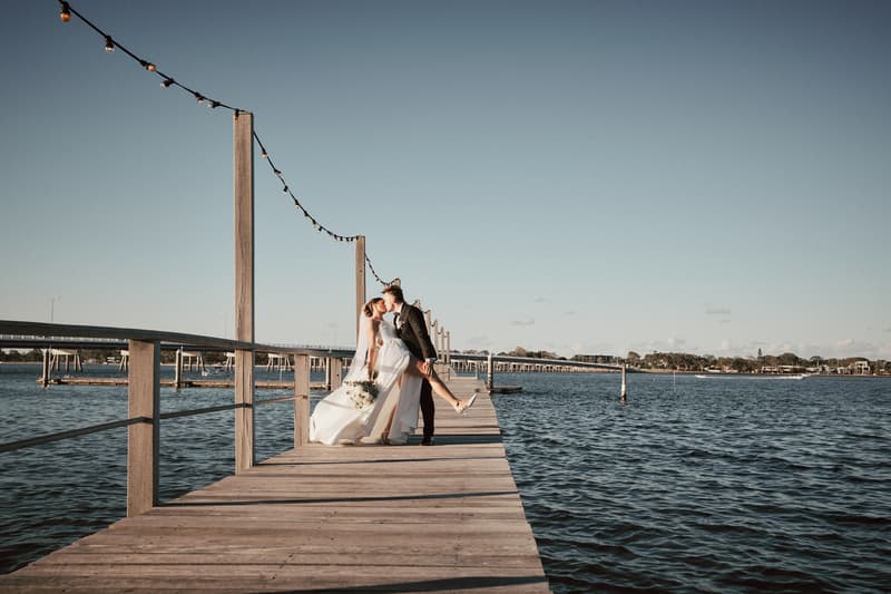 The bride Ashleigh and groom James share a kiss on a wooden pier at Sandstone Point Hotel, with the bride holding a bouquet and the water surrounding them under a clear sky.