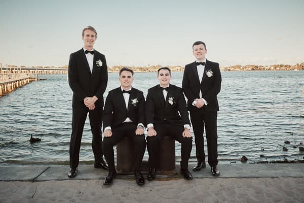 Four groomsmen in black tuxedos with white boutonnieres pose by the water, two seated on bollards and two standing behind them. Shortest day wedding timeline by She Said Yes.