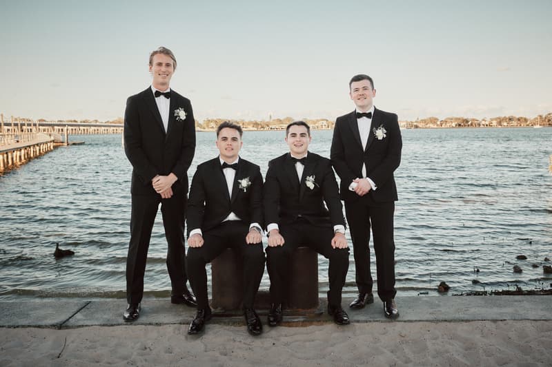 Four groomsmen in black tuxedos with white boutonnieres pose by the water at Sandstone Point Hotel, two seated on bollards and two standing behind them.