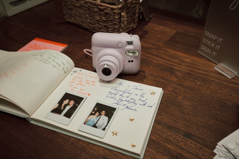 A wedding guestbook with handwritten messages and two instant photos sits on a wooden table at Sandstone Point Hotel — The Cellar, next to a white instant camera and a sign reading 'SNAP IT STICK IT SIGN IT'.