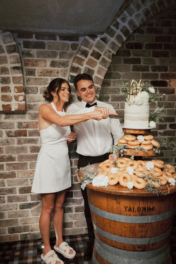 Bride Ashleigh and groom James cut their wedding cake, which is displayed on a wooden barrel decorated with donuts and flowers, at Sandstone Point Hotel — The Cellar.