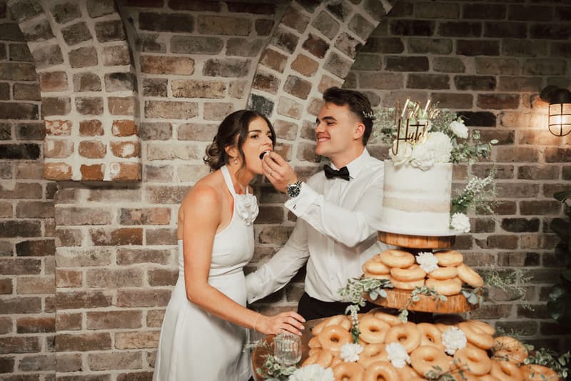 Bride Ashleigh and groom James at Sandstone Point Hotel — The Cellar reception stage, with James feeding Ashleigh a piece of cake next to a tiered cake and donut display.