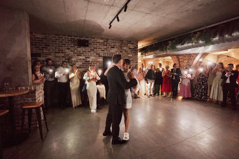 Bride Ashleigh and groom James share their first dance at the reception in The Cellar at Sandstone Point Hotel, surrounded by guests taking photos and watching.