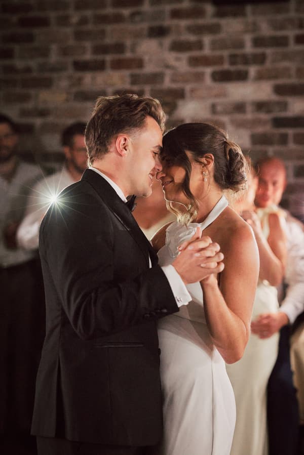 Bride Ashleigh and groom James share a close dance at the reception in Sandstone Point Hotel — The Cellar, with guests watching in the background against a brick wall.