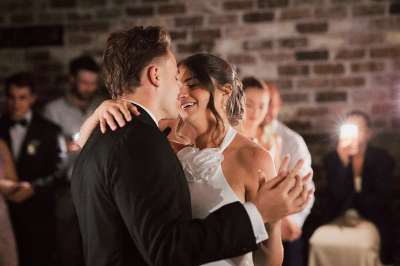Bride Ashleigh and groom James share a dance at the reception in The Cellar at Sandstone Point Hotel, with guests watching and one guest capturing the moment on a phone.